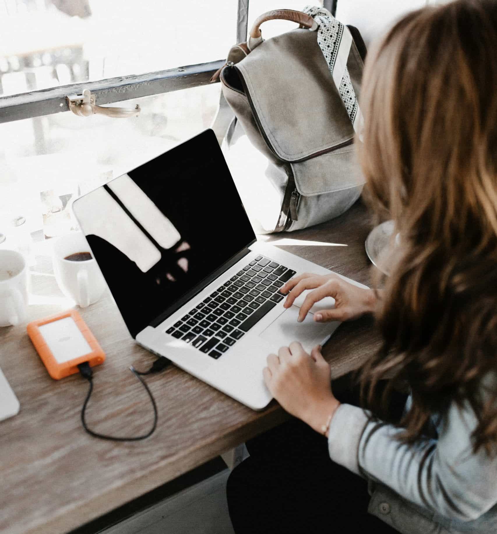 A woman is sat in the window seat of a cafe on her laptop with an orange hard drive to the side of her computer, plugged in. She's probably thinking about how she's going to be sky rocketing her social media growth using Quuu